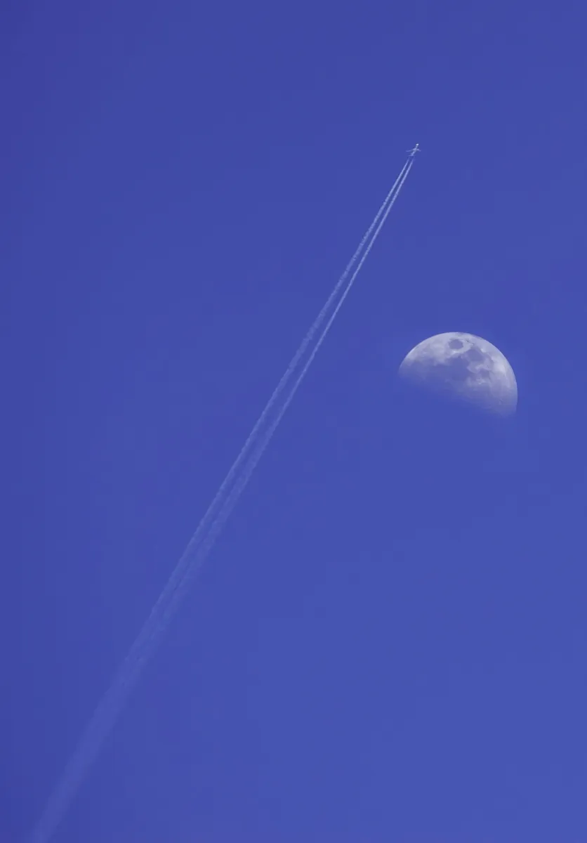 Daytime moon beside a white airplane contrail